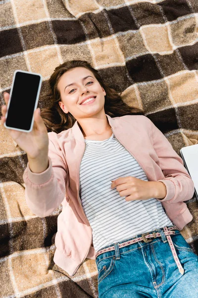 Vista aérea de la niña feliz sosteniendo el teléfono inteligente con pantalla en blanco mientras está acostado en una manta a cuadros - foto de stock