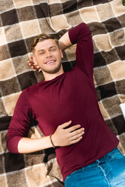 Blick von oben auf einen fröhlichen jungen Mann, der lächelnd auf einer karierten Decke liegt — Stockfoto