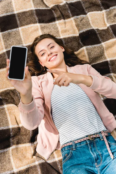Visão aérea da menina feliz apontando com o dedo para o smartphone com tela em branco enquanto deitado em cobertor quadriculado — Fotografia de Stock