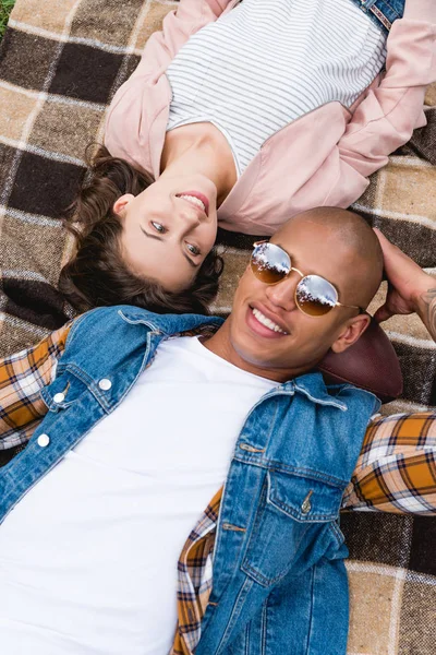 Overhead view of cheerful interracial couple lying on checkered blanket — Stock Photo