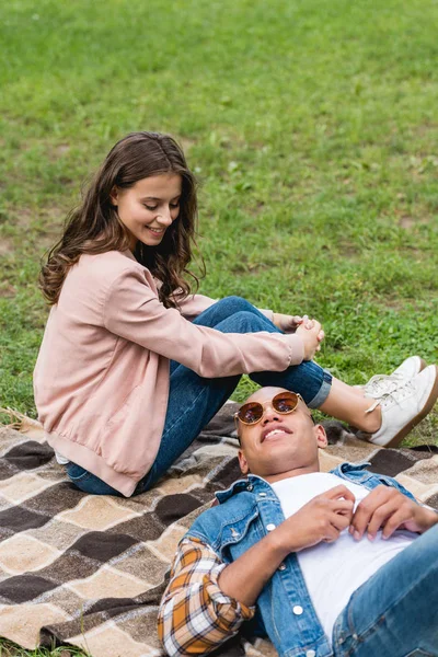 Attraente ragazza guardando africano americano fidanzato sdraiato su coperta in parco — Foto stock