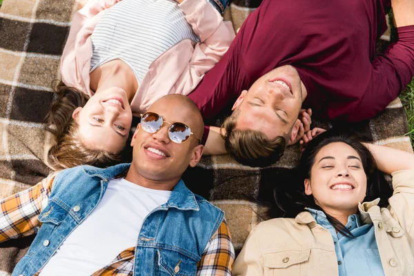 Blick von oben auf fröhliche multikulturelle Freunde, die auf karierter Decke liegen — Stockfoto