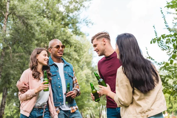 Amis multiculturels joyeux tenant des bouteilles avec de la bière et parlant dans le parc — Photo de stock