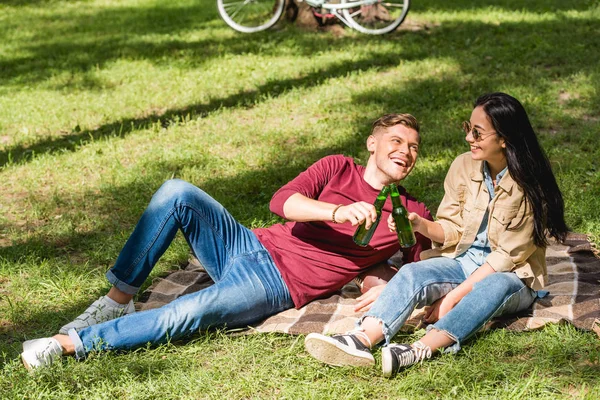 Feliz pareja tintineo botellas con cerveza mientras está sentado en manta en el parque - foto de stock
