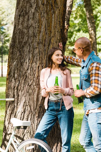 Fröhliches Mädchen schaut ihren afrikanisch-amerikanischen Freund an, während sie eine Flasche in der Hand hält und am Baum steht — Stockfoto