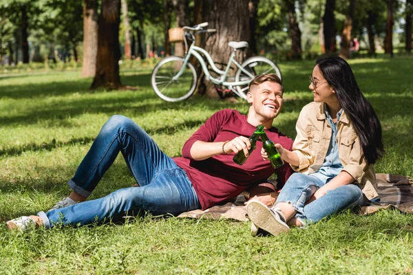 Cheerful couple clinking bottles with beer while sitting on blanket in park — Stock Photo