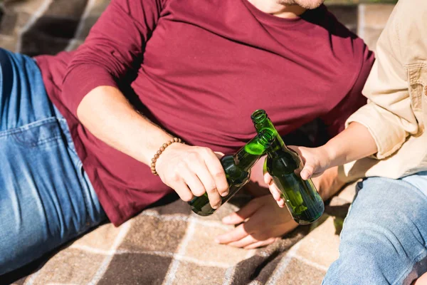 Cropped view of couple clinking bottles with beer while sitting on blanket — Stock Photo