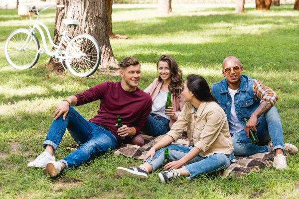 Багатоетнічна група друзів, що сидять на плетеній ковдрі і тримають пляшки в парку — стокове фото