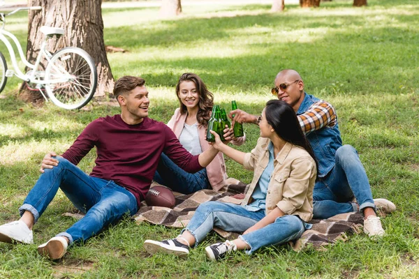 Gruppo multietnico di amici seduti su coperta a quadri e bottiglie di clinking nel parco — Foto stock