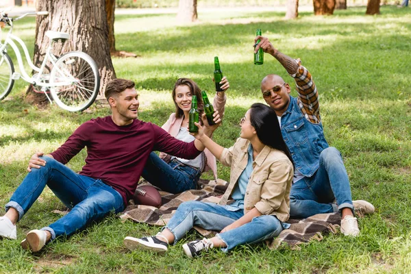 Feliz grupo multiétnico de amigos sentados en manta a cuadros y botellas tintineo en el parque - foto de stock