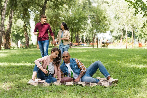 Selective focus of cheerful african american young man and attractive girl sitting on blanket and holding bottles of beer with friends on background — Stock Photo