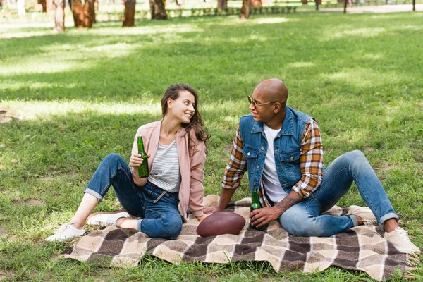 Happy interracial couple looking at each other while sitting on blanket with bottles of beer — Stock Photo