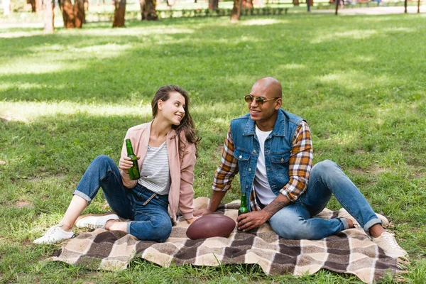 Menina feliz olhando para o namorado americano africano enquanto sentado em cobertor com garrafa de cerveja — Fotografia de Stock