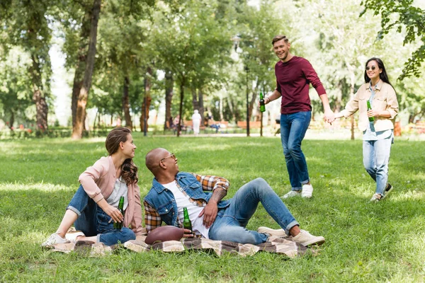 African american young man and happy girl sitting on blanket with bottles of beer and looking at friends holding hands — Stock Photo