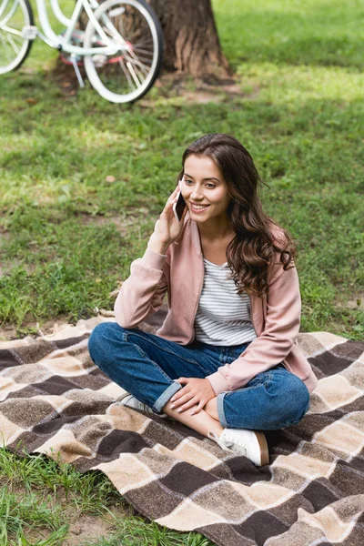 Ragazza allegra parlando su smartphone mentre seduto su una coperta a scacchi nel parco — Foto stock