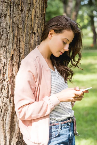 Привлекательная девушка смотрит на смартфон, стоя рядом с деревом в парке — стоковое фото