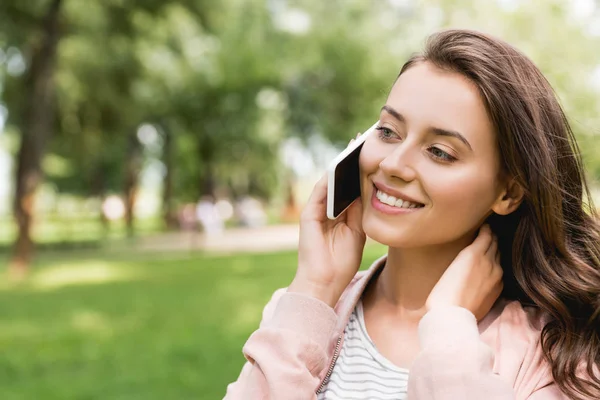 Attraktives Mädchen, das auf dem Smartphone spricht und im Park lächelt — Stockfoto