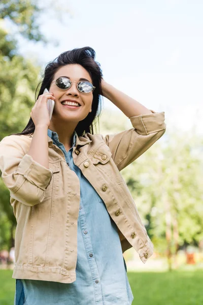 Fröhliche junge Frau mit Sonnenbrille spricht im Park mit Smartphone — Stockfoto