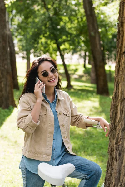 Щаслива молода жінка в сонцезахисних окулярах говорить на смартфоні біля велосипеда в парку — стокове фото