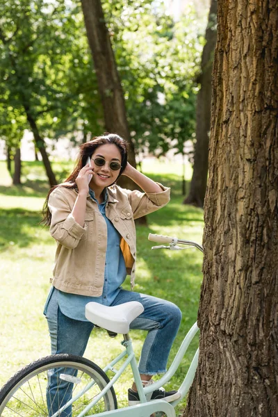 Fröhliche junge Frau mit Sonnenbrille unterhält sich mit Smartphone in der Nähe von Fahrrad im Park — Stockfoto