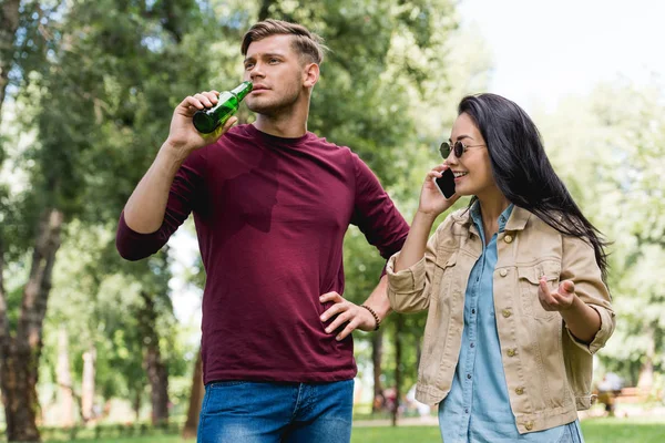 Gaie fille dans des lunettes de soleil parler sur smartphone près petit ami boire de la bière dans le parc — Photo de stock