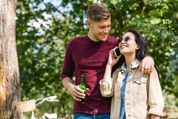 Alegre chica en gafas de sol hablando en smartphone cerca novio celebración botella de cerveza en parque - foto de stock