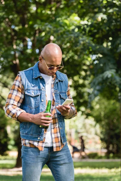 Bonito afro-americano homem olhando para smartphone enquanto segurando garrafa — Fotografia de Stock