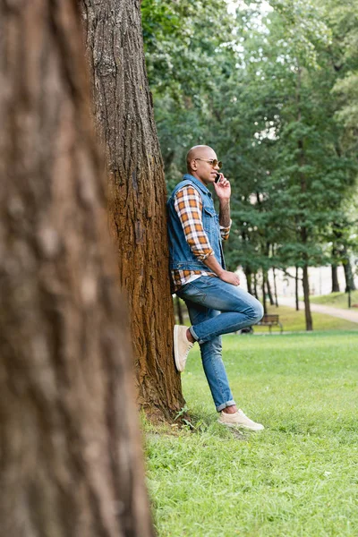 Избирательный фокус красивого африканского американца, разговаривающего по смартфону, стоя в парке — стоковое фото