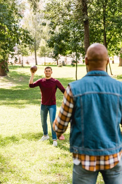 Back view of african american man looking at happy friend throwing american football in park — Stock Photo