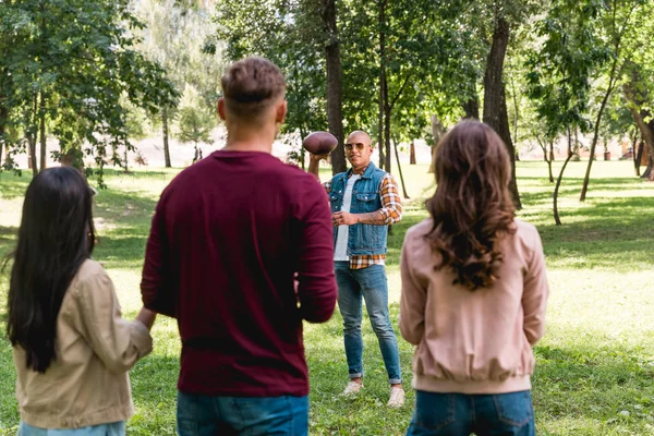 Back view of friends looking at african american man holding american football in park — Stock Photo