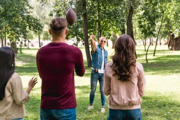 Visão traseira dos amigos olhando para o homem americano africano jogando futebol americano no parque — Fotografia de Stock