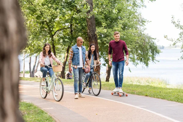 Afro-americano homem andando perto de meninas andar de bicicleta e alegre amigo longboarding no parque — Fotografia de Stock