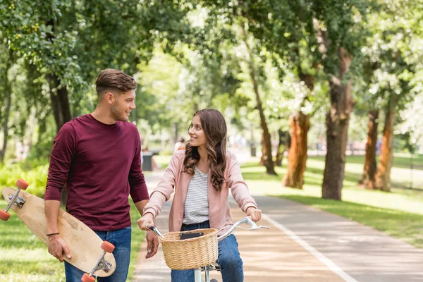Handsome man holding longboard and looking at attractive girlfriend riding bicycle — Stock Photo