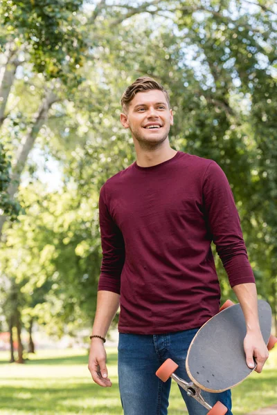 Bello giovane uomo sorridente mentre tiene longboard nel parco — Foto stock