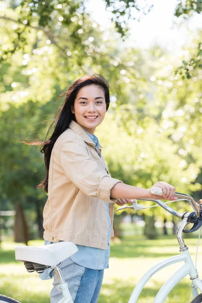 Счастливая симпатичная девушка улыбается, держа велосипед в парке — стоковое фото