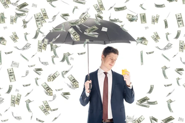 Exitoso hombre de negocios en traje con paraguas y alcancía amarilla bajo la lluvia de dinero sobre fondo blanco - foto de stock