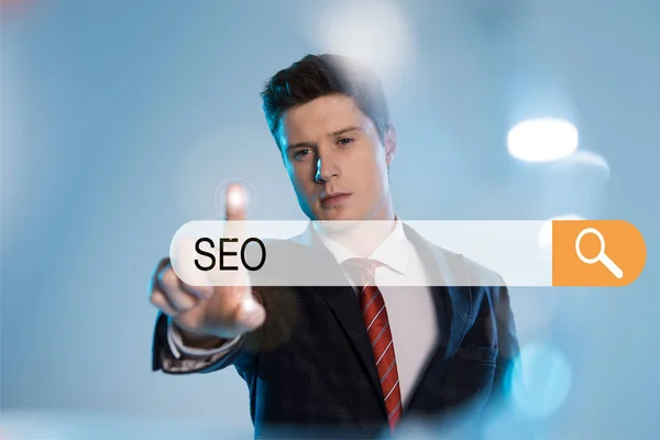 Confident businessman in suit pointing with finger at search bar with seo letters in front on blue background — Stock Photo