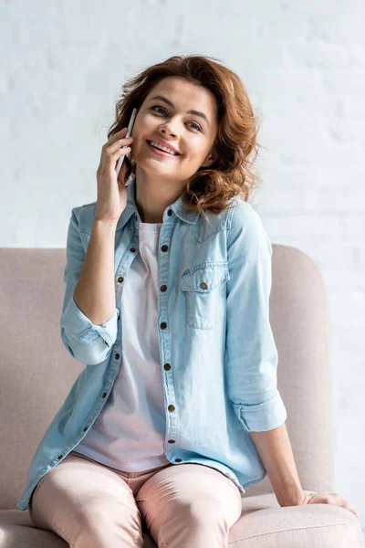 Mujer feliz sentada en el sofá y hablando en el teléfono inteligente con sonrisa en gris - foto de stock