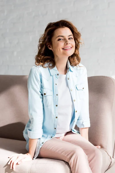 Blissful curly woman in shirt sitting on sofa and smiling on grey — Stock Photo