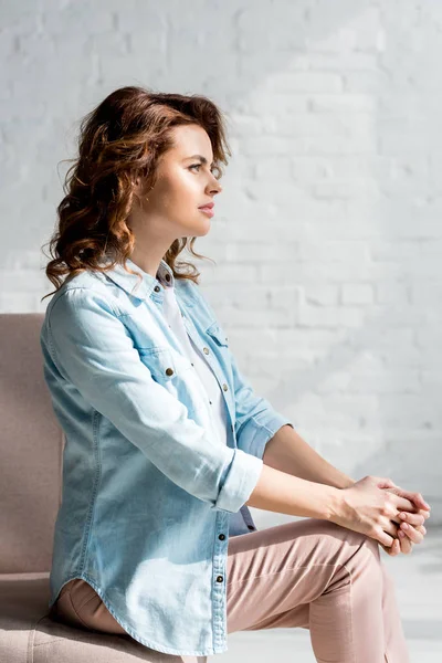 Beautiful curly woman in shirt sitting on sofa on grey — Stock Photo