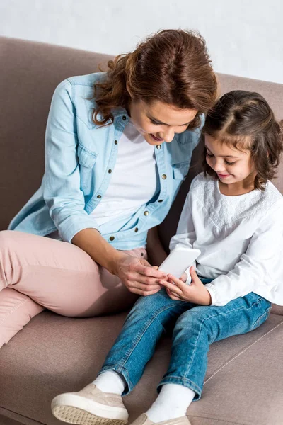 Cheerful mother and daughter sitting on brown sofa and using smartphone isolated on grey — Stock Photo