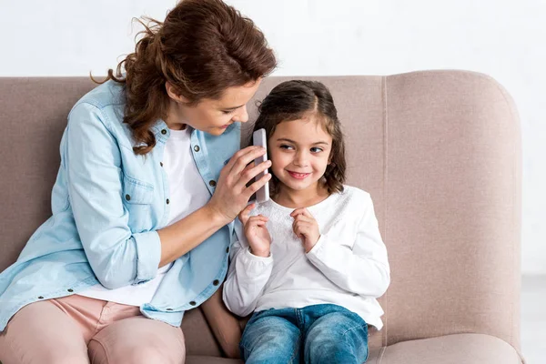 Madre e hija sentadas en un sofá marrón y hablando en un smartphone en blanco - foto de stock