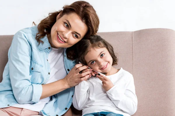 Blissful mother and daughter sitting on brown sofa and talking on smartphone isolated on white — Stock Photo