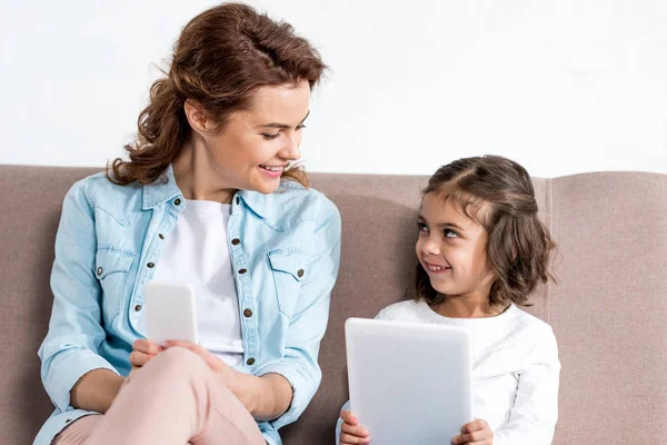 Mãe com smartphone e filha com tablet digital sentados no sofá e olhando uns para os outros isolados no branco — Fotografia de Stock