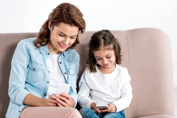 Mère et fille heureuses assises sur un canapé marron et utilisant des smartphones sur blanc — Photo de stock