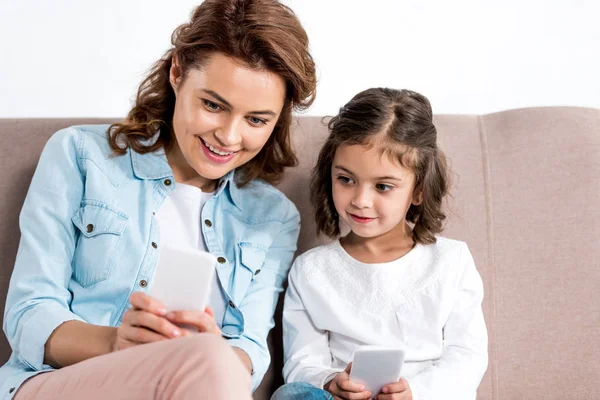 Alegre madre e hija sentadas en el sofá y usando teléfonos inteligentes aislados en blanco - foto de stock