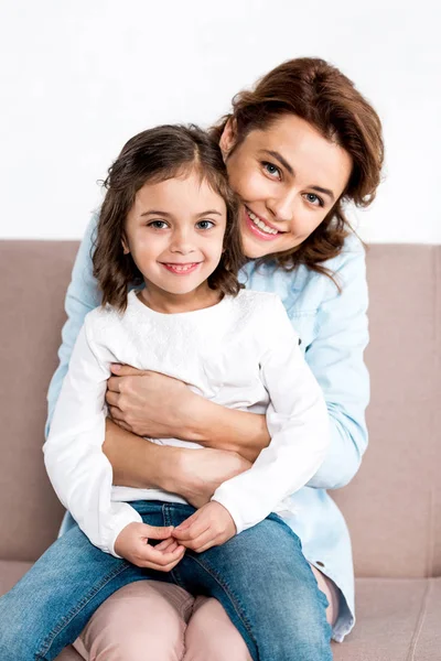 Rindo mãe sentada no sofá e segurando filha de joelhos isolados em branco — Fotografia de Stock