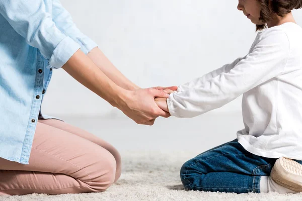 Vista ritagliata di madre e figlia sedute su tappeto e che si tengono per mano sul bianco — Foto stock