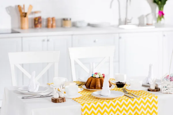 Osterkuchen mit bemalten Eiern, Keramikhasen und Geschirr auf dem Tisch in der Küche — Stockfoto