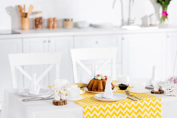 Gâteau de Pâques avec œufs peints, lapins en céramique et vaisselle sur la table dans la cuisine — Photo de stock